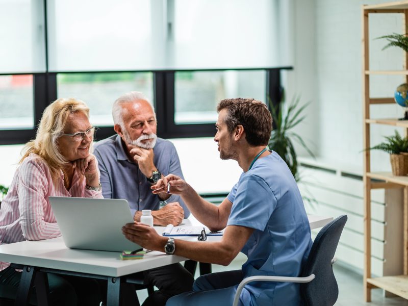 Happy mature couple and their doctor communicating while using laptop during medical appointment.